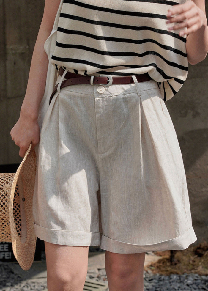 Frau in gestreiftem Oberteil und beigen Leinen-Shorts mit braunem Gürtel, hält Strohhut. Sommermode, lässiger Stil, trendige Damenbekleidung.