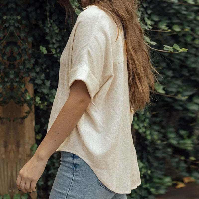 Frau in beiger Bluse und blauer Jeans vor grüner Pflanzenwand. Lässige Mode, Sommeroutfit, Naturhintergrund.