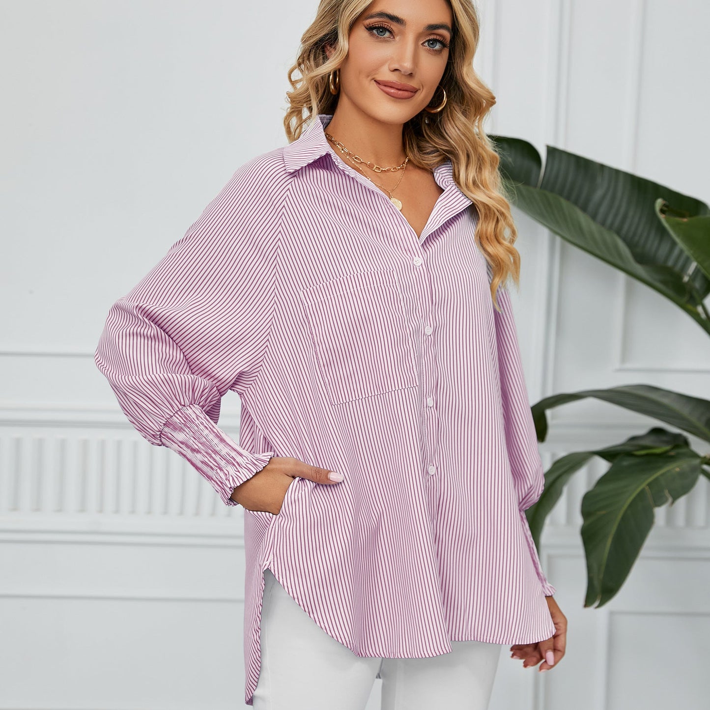 Frau in gestreifter Bluse, lächelnd, mit Hand in der Tasche, vor weißer Wand und Pflanze. Modetrend, Damenmode, Freizeitlook.