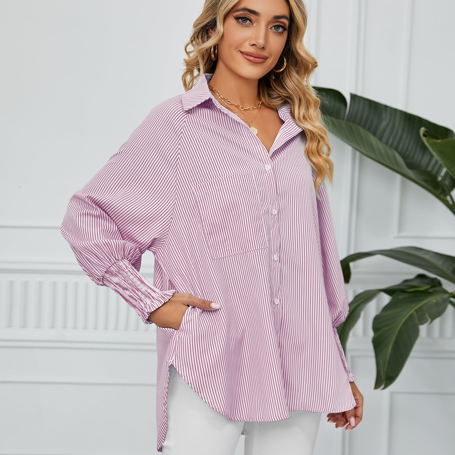 Frau in gestreifter Bluse, lächelnd, mit Hand in der Tasche, vor weißer Wand und Pflanze. Modetrend, Damenmode, Freizeitlook.