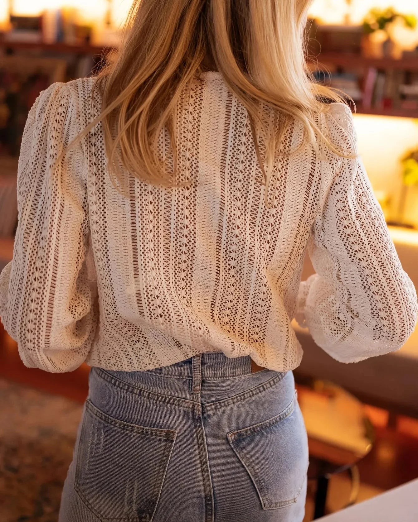 Frau in weißer Spitzenbluse und blauer Jeans von hinten, stilvolle Mode, gemütliche Innenraumbeleuchtung, trendige Damenbekleidung.