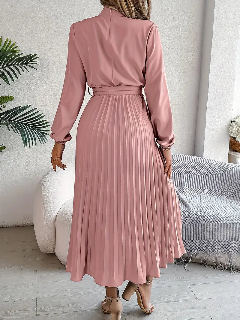 Frau in rosa, langem, plissiertem Kleid mit Gürtel, steht in modernem Wohnzimmer. Elegantes Mode-Outfit, ideal für stilvolle Anlässe.