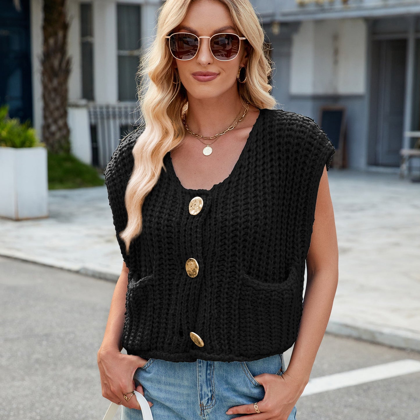 Frau mit blonder Welle, Sonnenbrille, schwarzer Strickweste mit großen Knöpfen, blauer Jeans, lässiger Straßenlook, modisch, urbaner Stil.