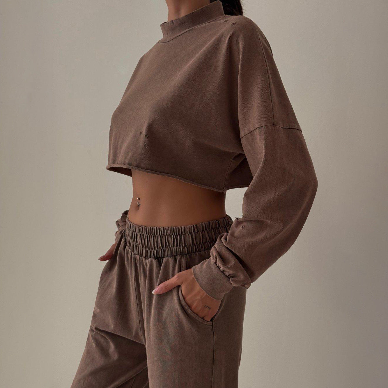 Frau in braunem, bauchfreiem Sweatshirt und Jogginghose, seitlich stehend. Modetrend, Freizeitkleidung, bequeme Damenmode.