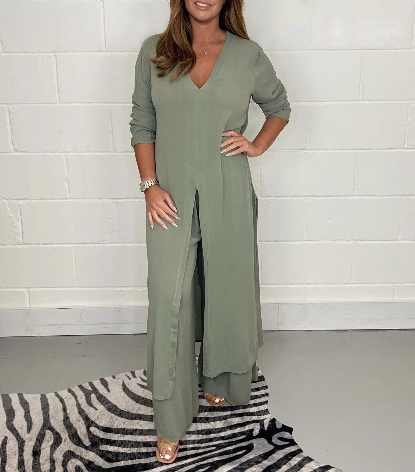 Frau in olivgrünem, langem Kleid mit V-Ausschnitt und Schlitz, steht auf grauem Boden mit Zebrateppich. Elegante Mode, lässiger Stil.