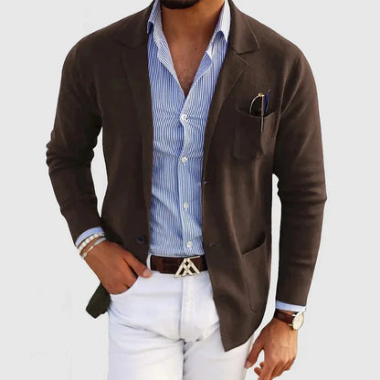 Mann in braunem Blazer, blau-weiß gestreiftem Hemd und weißer Hose, modisch elegant gekleidet, mit Armbändern und Uhr, vor grauem Hintergrund.