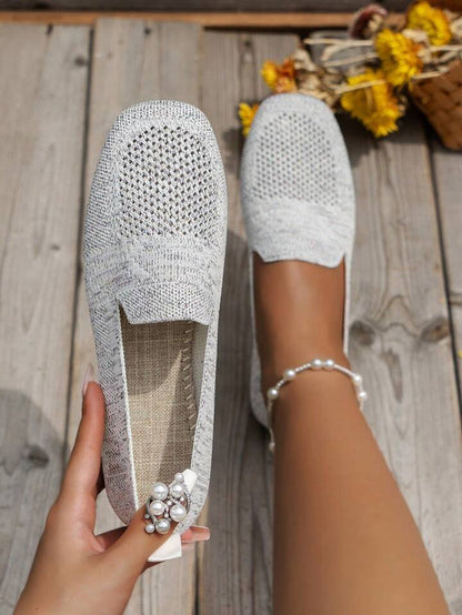 Damenhand hält grauen Strick-Slipper, getragen auf Holzterrasse, mit Perlenschmuck am Finger. Sommermode, bequeme Schuhe, Freizeitlook.
