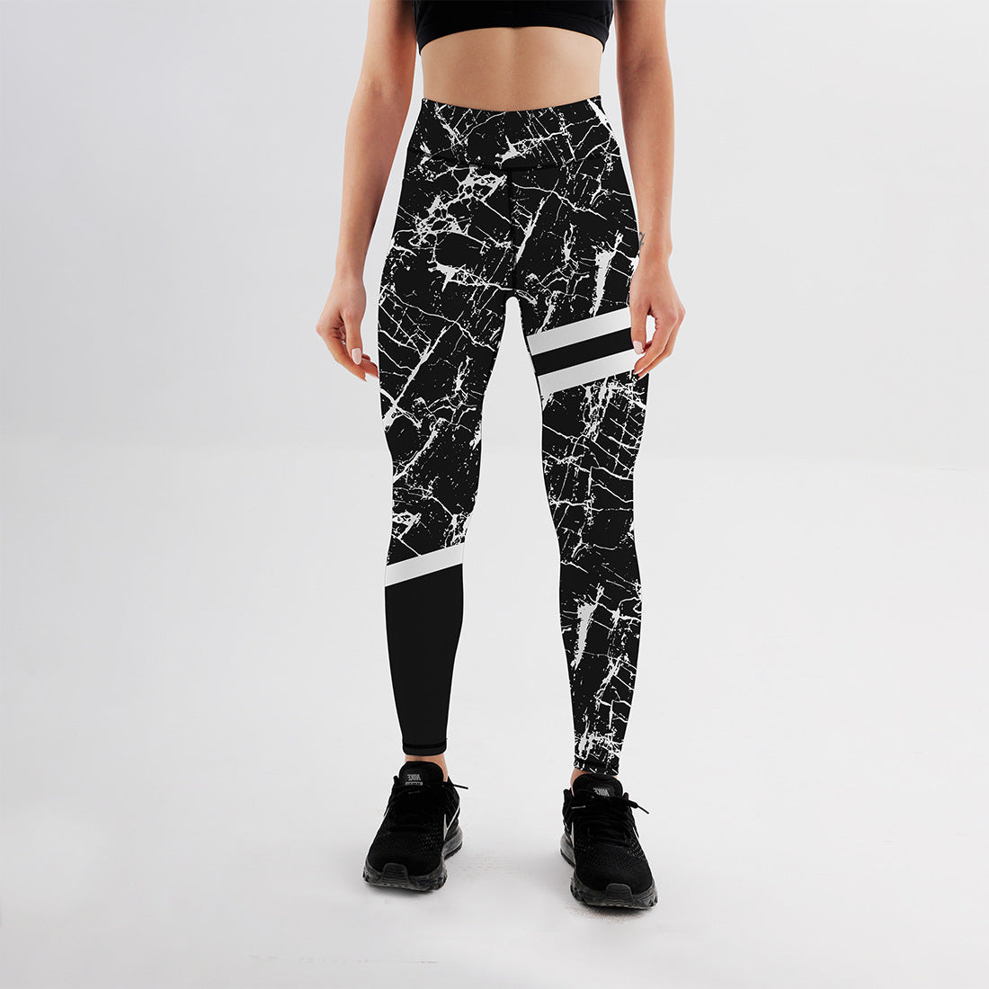 Frau in schwarzem Sport-BH und gemusterten Leggings, steht auf weißem Hintergrund. Fitnessmode, Sportbekleidung, Damenleggings, Athleisure.