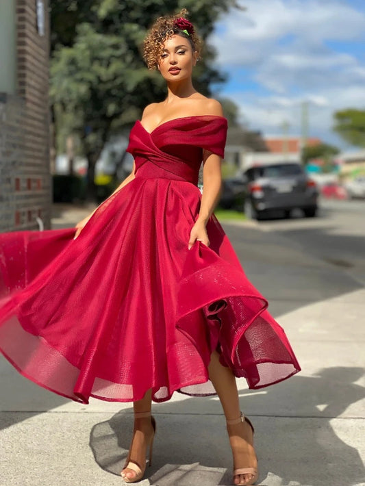 Frau in elegantem, schulterfreiem roten Kleid auf Gehweg, sommerlicher Hintergrund, Mode, stilvoll, urban, Tageslicht, schicke Abendgarderobe.