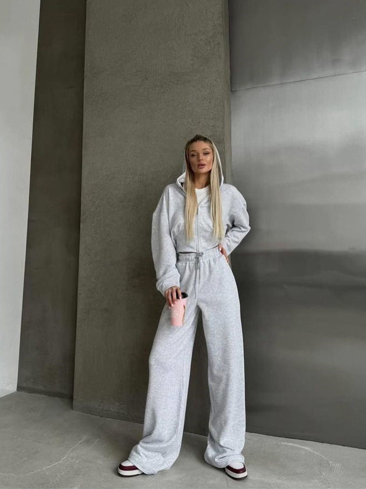 Graues Damen-Sweatshirt und Jogginghose, lässiger Look, Frau mit blonden Haaren, hält rosa Trinkflasche, moderne Betonwand im Hintergrund.