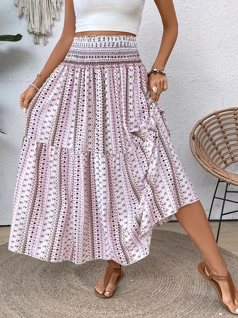 Frau in weißem Crop-Top und langem, geblümtem Boho-Rock, steht neben einem Rattan-Stuhl. Sommermode, Boho-Stil, Freizeitkleidung.