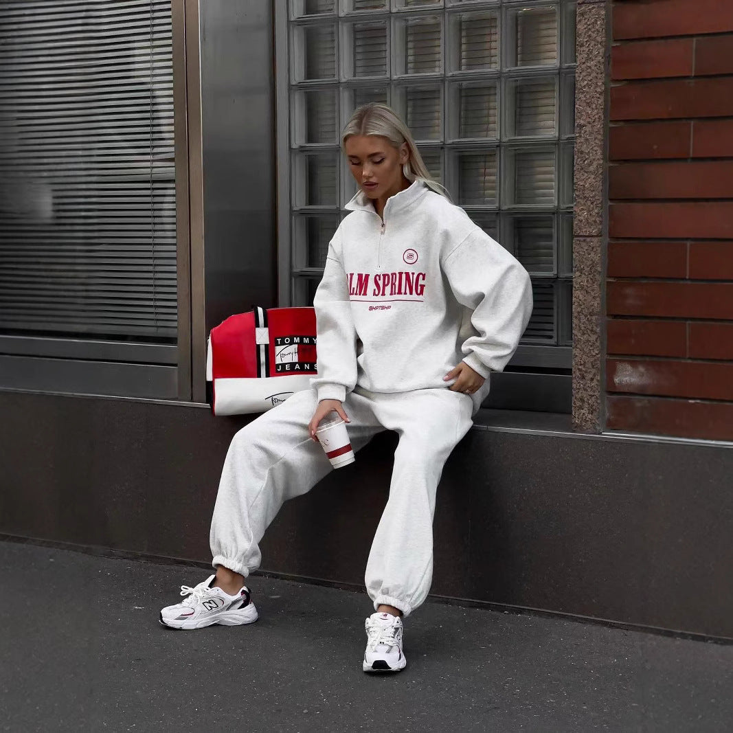Frau in weißem Jogginganzug mit "Palm Spring" Aufdruck, sitzt auf einer Bank. Neben ihr eine Tommy Hilfiger Tasche. Trägt weiße Sneakers.