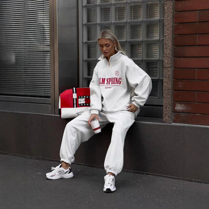 Frau in weißem Jogginganzug mit "Palm Spring" Aufdruck, sitzt auf einer Bank. Neben ihr eine Tommy Hilfiger Tasche. Trägt weiße Sneakers.