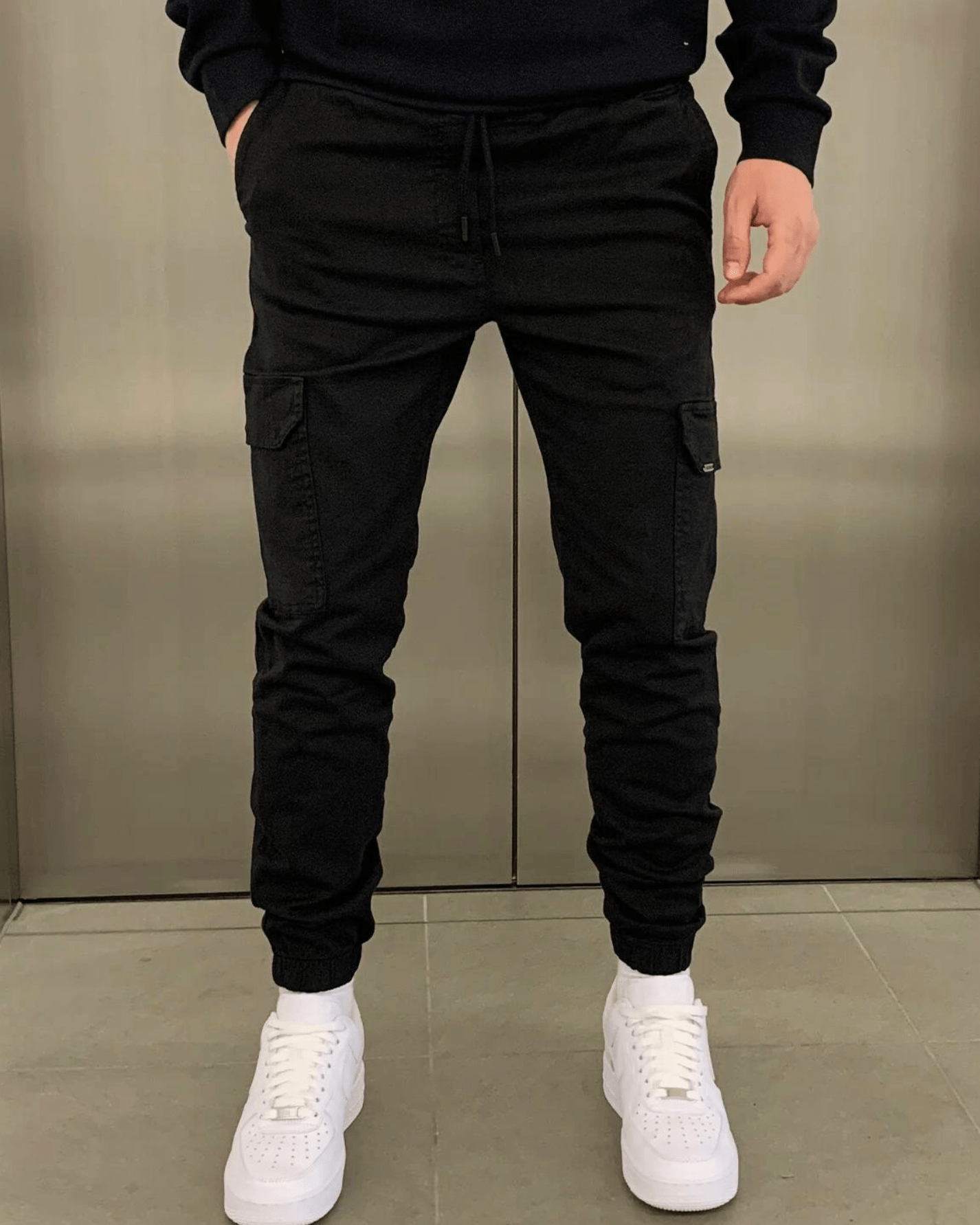Mann in schwarzer Cargohose und weißen Sneakers vor grauem Hintergrund. Streetwear, Herrenmode, lässiger Stil.