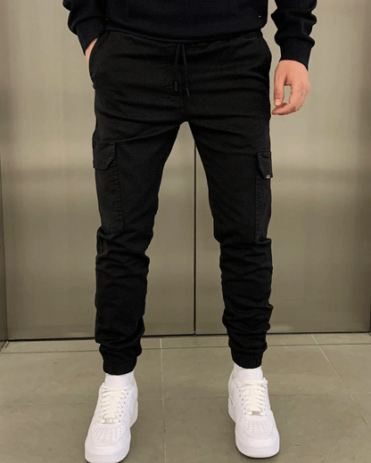 Mann in schwarzer Cargohose und weißen Sneakers vor grauem Hintergrund. Streetwear, Herrenmode, lässiger Stil.