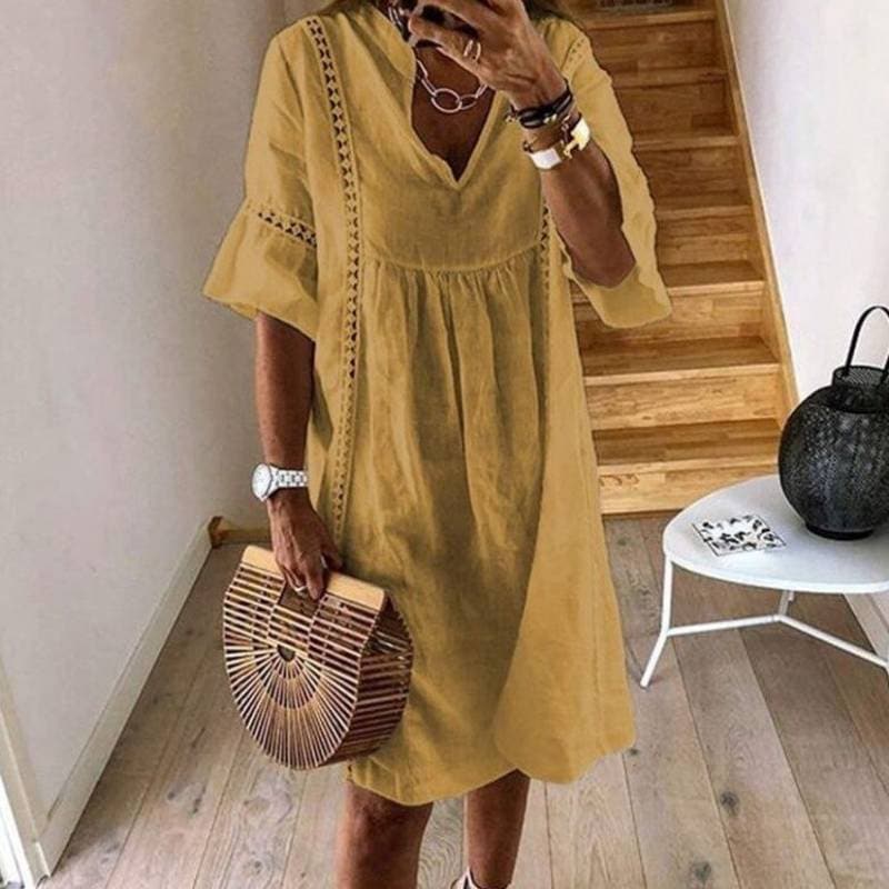 Frau in gelbem Sommerkleid mit V-Ausschnitt, hält eine Bambus-Handtasche. Steht vor einer Holztreppe, trägt Armbänder und eine Uhr.