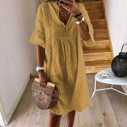 Frau in gelbem Sommerkleid mit V-Ausschnitt, hält eine Bambus-Handtasche. Steht vor einer Holztreppe, trägt Armbänder und eine Uhr.