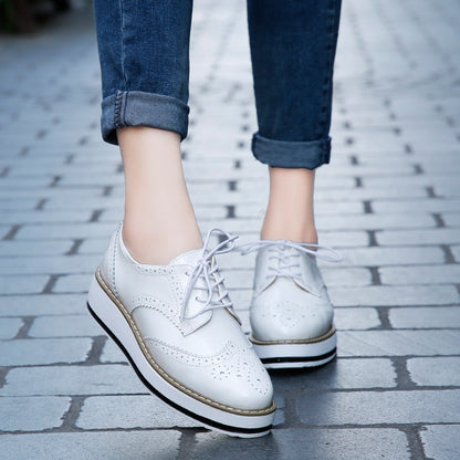 Weiße Brogues-Schuhe mit Plateau, getragen von einer Person in hochgekrempelten Jeans, auf einem gepflasterten Gehweg. Modische Damen-Sneaker.