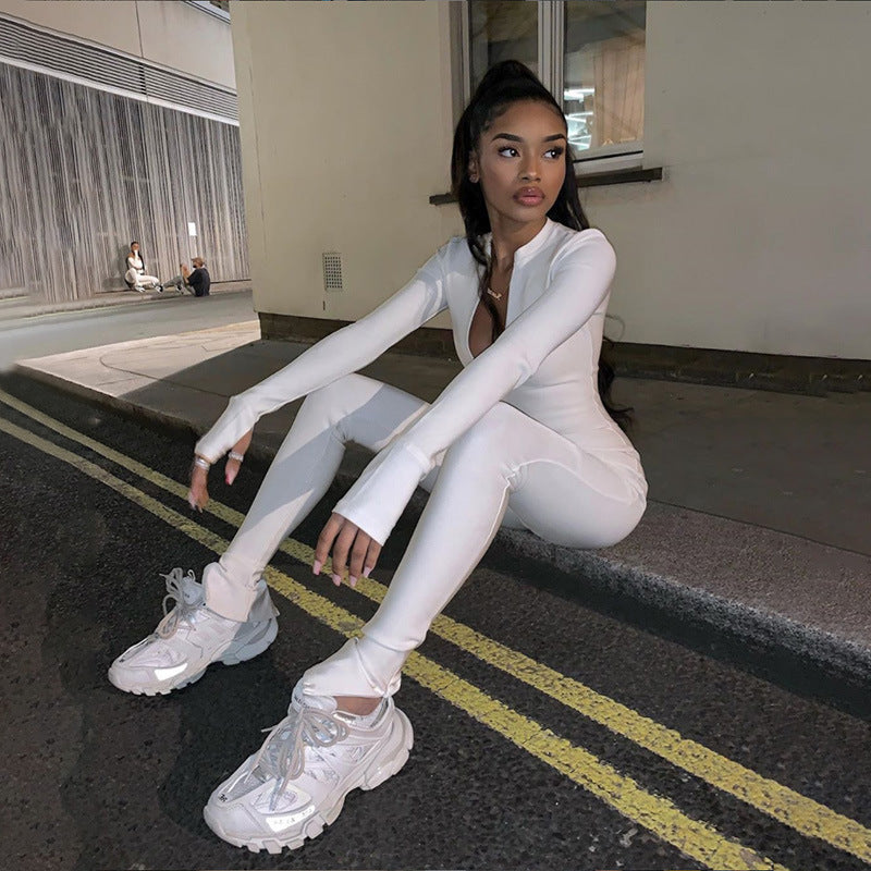 Frau in weißem Jumpsuit und Sneakers sitzt auf Bordstein in urbaner Umgebung bei Nacht. Streetwear, Mode, Nachtfotografie.