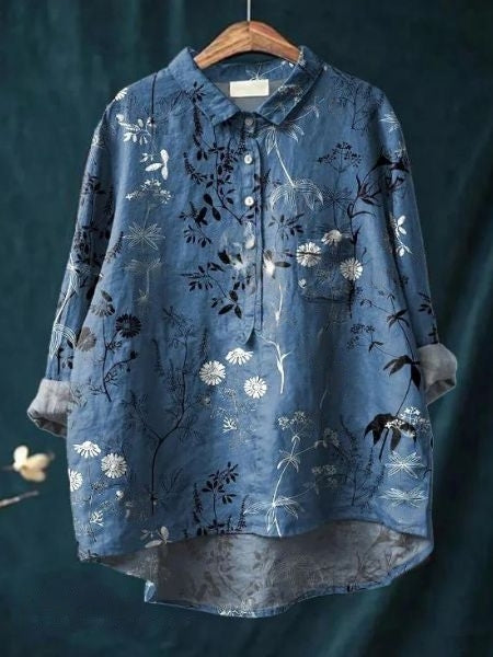 Blaues Hemd mit floralen Mustern, langen Ärmeln und Knopfleiste, auf einem Holzkleiderbügel vor dunkelgrünem Hintergrund. Elegante Damenmode.