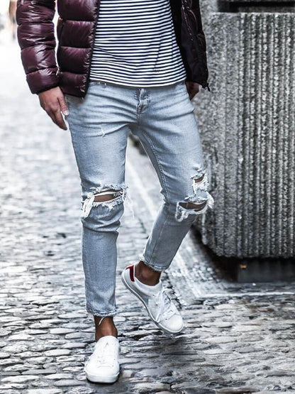 Mann in zerrissenen Jeans, gestreiftem Shirt und Daunenjacke, trägt weiße Sneaker. Modischer Streetstyle-Look auf Kopfsteinpflaster.
