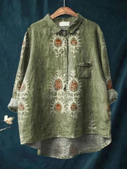 Grünes Leinenhemd mit floralen Mustern, Knopfleiste und Brusttasche, hängend auf Holzkleiderbügel vor blauem Hintergrund. Boho-Stil, Damenmode.