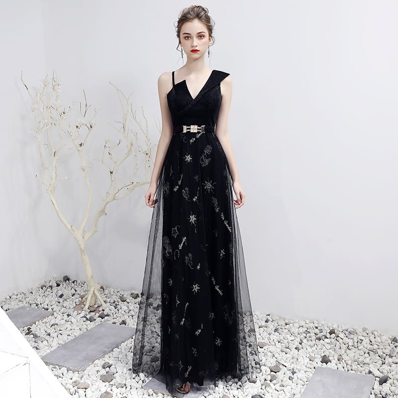Elegantes schwarzes Abendkleid mit V-Ausschnitt und Gürtel, verziert mit funkelnden Details, auf Kiesboden vor weißer Wand und kahlem Baum.