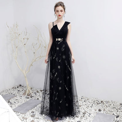 Elegantes schwarzes Abendkleid mit V-Ausschnitt und Gürtel, verziert mit funkelnden Details, auf Kiesboden vor weißer Wand und kahlem Baum.