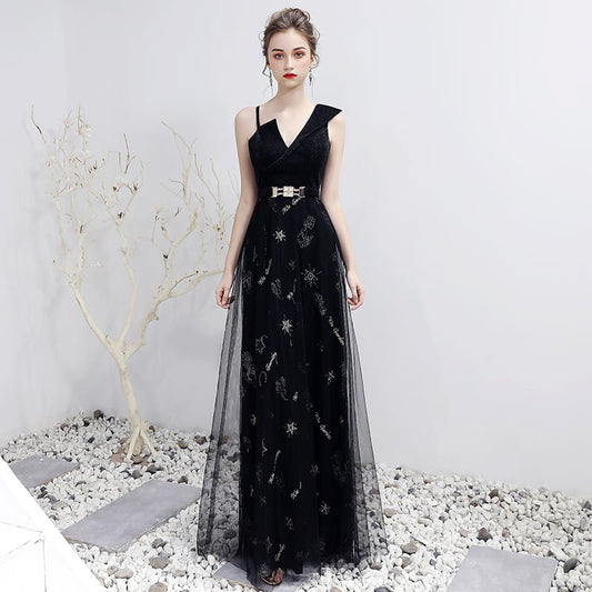 Elegantes schwarzes Abendkleid mit V-Ausschnitt und Gürtel, verziert mit funkelnden Details, auf Kiesboden vor weißer Wand und kahlem Baum.