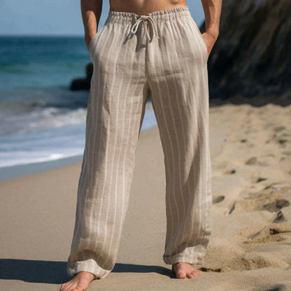 Person in beigen Leinenhose am Strand, barfuß im Sand, mit Meer im Hintergrund. Sommermode, Strandkleidung, entspannter Stil.