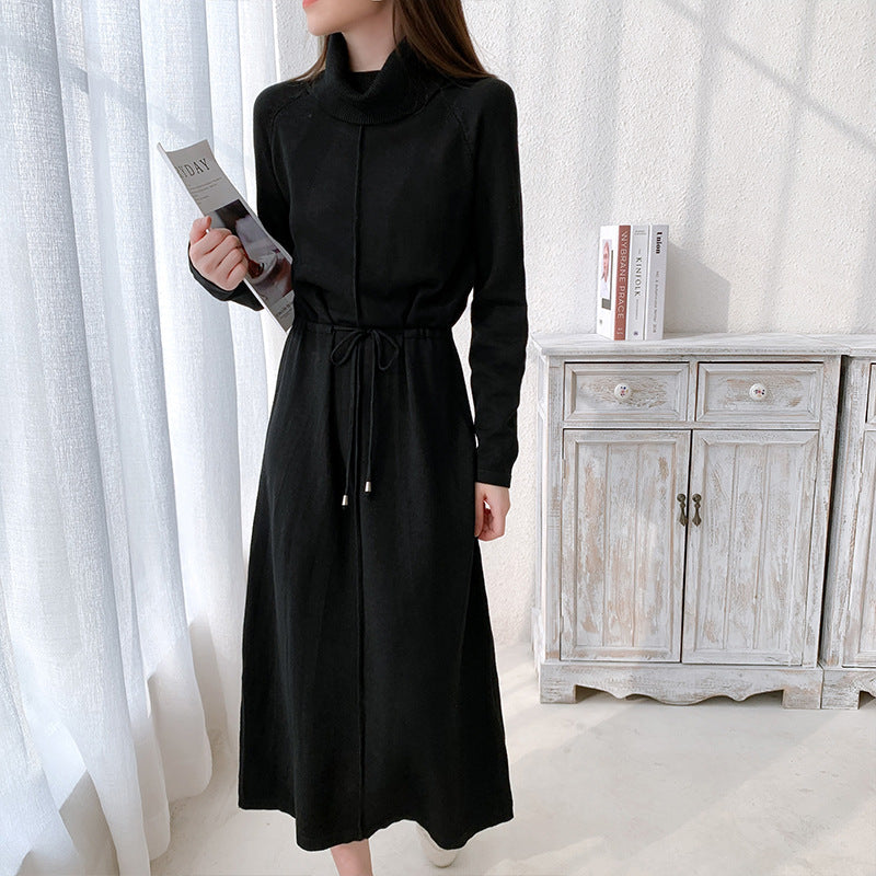Frau in elegantem, schwarzem Strickkleid mit Stehkragen, hält Zeitschrift. Stehendes Möbelstück im Hintergrund. Modetrend, Damenmode, Herbstkleid.