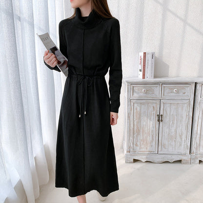 Frau in elegantem, schwarzem Strickkleid mit Stehkragen, hält Zeitschrift. Stehendes Möbelstück im Hintergrund. Modetrend, Damenmode, Herbstkleid.