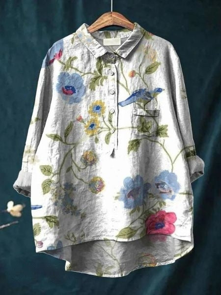 Graues Leinenhemd mit buntem Blumen- und Vogelmuster, hängend auf einem Holzkleiderbügel vor dunkelblauem Hintergrund.
