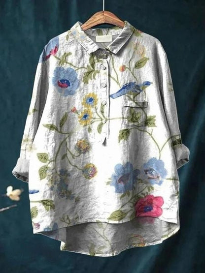 Graues Leinenhemd mit buntem Blumen- und Vogelmuster, hängend auf einem Holzkleiderbügel vor dunkelblauem Hintergrund.
