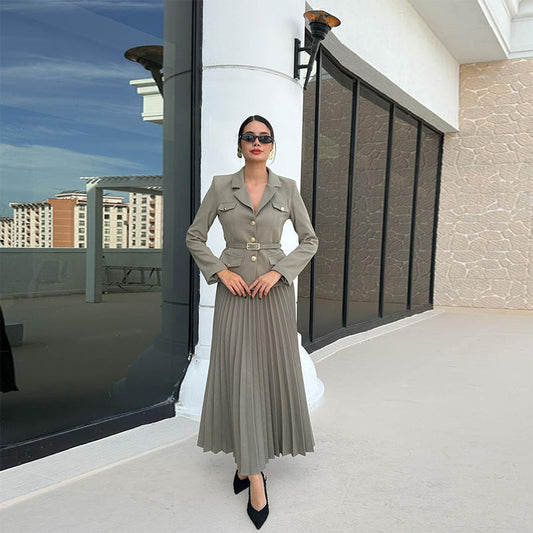 Elegante Frau in grauem Blazer und Faltenrock, steht vor modernem Gebäude mit Glasfassade. Stilvolle Mode, Business-Look, urbane Architektur.