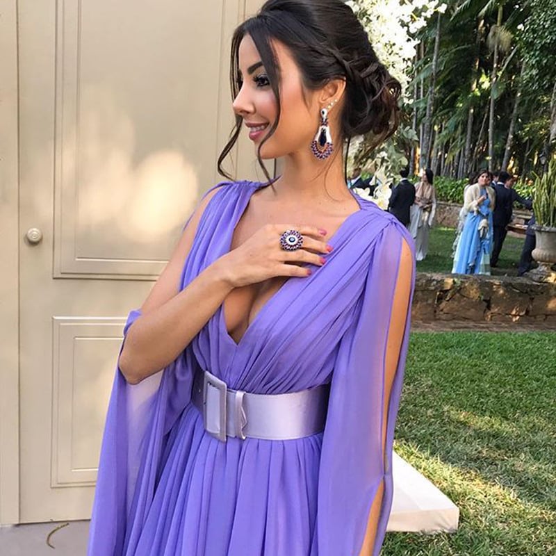 Eine Frau in einem eleganten, lila Abendkleid mit tiefem Ausschnitt und silbernem Gürtel, trägt auffälligen Schmuck, steht im Freien.