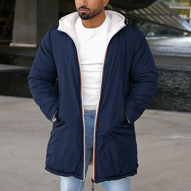 Mann in blauem Wintermantel mit Kapuze, weißem Pullover und Jeans steht draußen. Moderne Architektur im Hintergrund. Wintermode, Herrenbekleidung.