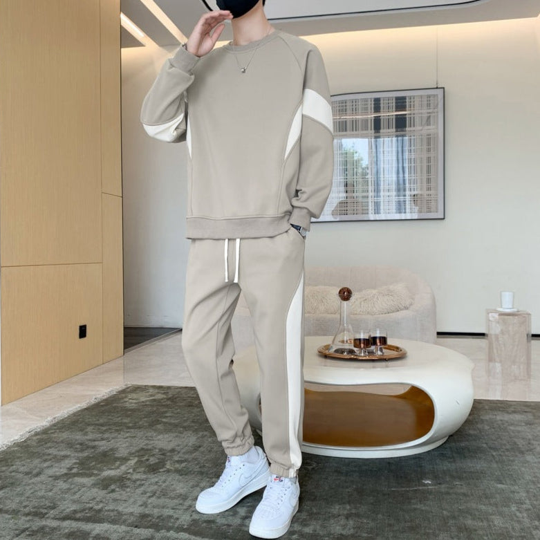 Person in beigem Trainingsanzug mit weißen Streifen steht in modernem Wohnzimmer. Stilvolle Freizeitmode, weiße Sneakers, minimalistisches Interieur.