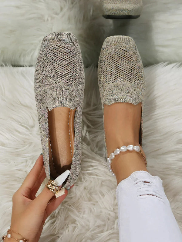 Damenhand hält grauen Strick-Loafer auf weißem Teppich, getragen mit zerrissener weißer Jeans und Perlenarmband. Bequeme, stilvolle Schuhe.