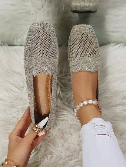 Damenhand hält grauen Strick-Loafer auf weißem Teppich, getragen mit zerrissener weißer Jeans und Perlenarmband. Bequeme, stilvolle Schuhe.