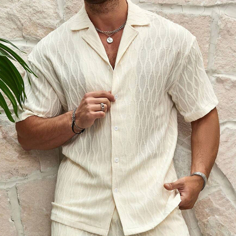 Mann in cremefarbenem, kurzärmligem Hemd mit Knopfleiste und Ketten vor Steinwand. Sommermode, Herrenbekleidung, lässiger Stil.