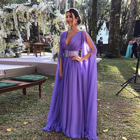 Frau in fliederfarbenem Abendkleid mit tiefem Ausschnitt und Umhang-Ärmeln, steht im Freien bei einer Hochzeit, umgeben von Bäumen und Blumen.