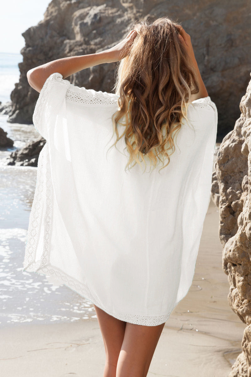 Frau am Strand in weißem Strandkleid, Rückenansicht, blonde Haare, felsige Küste, Sommermode, entspannte Atmosphäre, Urlaub am Meer.