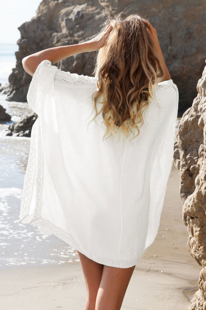 Frau am Strand in weißem Strandkleid, Rückenansicht, blonde Haare, felsige Küste, Sommermode, entspannte Atmosphäre, Urlaub am Meer.