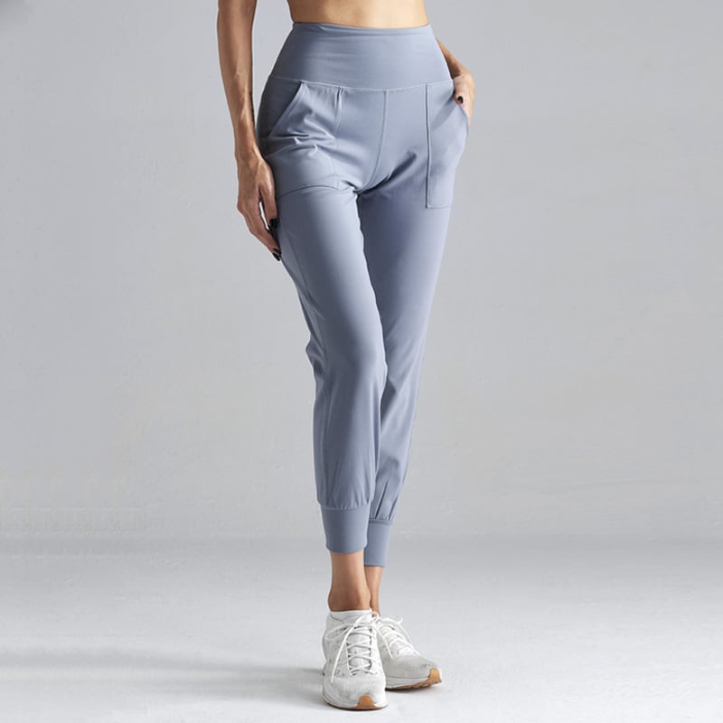 Damen-Sporthose in Hellgrau, hoher Bund, seitliche Taschen, knöchellang, kombiniert mit weißen Sneakers. Ideal für Fitness und Freizeit.