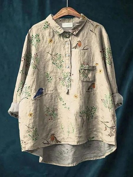 Beige Leinenbluse mit Vogel- und Pflanzenmuster, Knopfleiste und Brusttasche, hängend auf Holzkleiderbügel vor blauem Hintergrund.