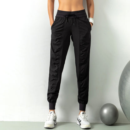 Frau in schwarzen Jogginghosen und weißen Sneakers vor grauer Wand, Fitnessball daneben. Sportliche Kleidung, Freizeitmode, Damenbekleidung.