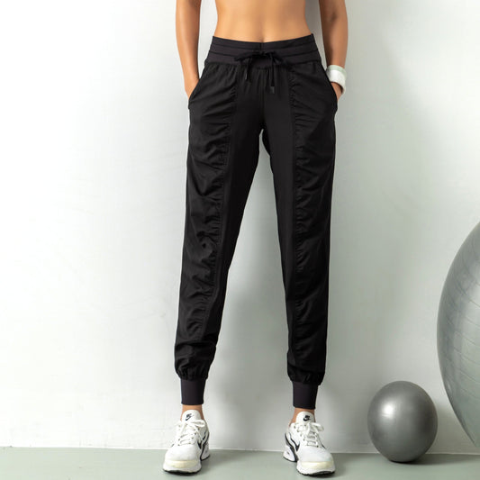 Frau in schwarzen Jogginghosen und weißen Sneakers vor grauer Wand, Fitnessball daneben. Sportliche Kleidung, Freizeitmode, Damenbekleidung.