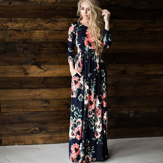 Blonde Frau in langem, geblümtem Kleid vor Holzwand. Elegantes Maxikleid mit floralen Mustern, ideal für Mode und Stil.