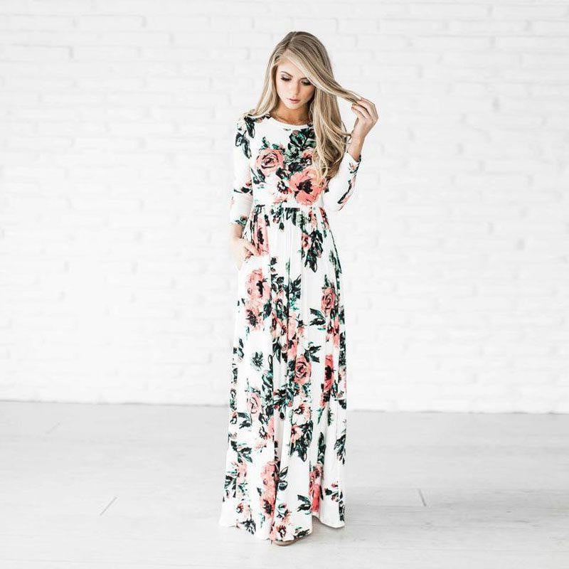 Frau in langem, geblümtem Kleid vor weißer Backsteinwand, modisches Outfit, florales Design, eleganter Look, Damenmode, stilvolle Kleidung.